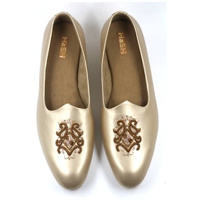 May include: A pair of gold-colored embroidered flat shoes with a brown embroidered design. The shoes have a pointed toe and a slip-on style. The brand name "Hosh" is printed on the inside of the shoes.