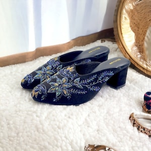 May include: A pair of black heeled mules with a floral embroidery design featuring iridescent sequins and gold thread. The shoes have a low block heel and a pointed toe.