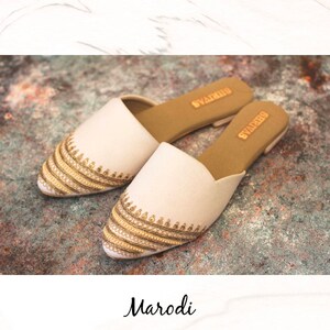 May include: A pair of white embroidered flat shoes with gold and beige embellishments. The shoes have a pointed toe and a slip-on design. The brand name "Swarovski" is visible on the insole.