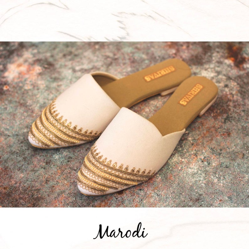 May include: A pair of white embroidered flat shoes with gold and beige embellishments. The shoes have a pointed toe and a slip-on design. The brand name "Swarovski" is visible on the insole.