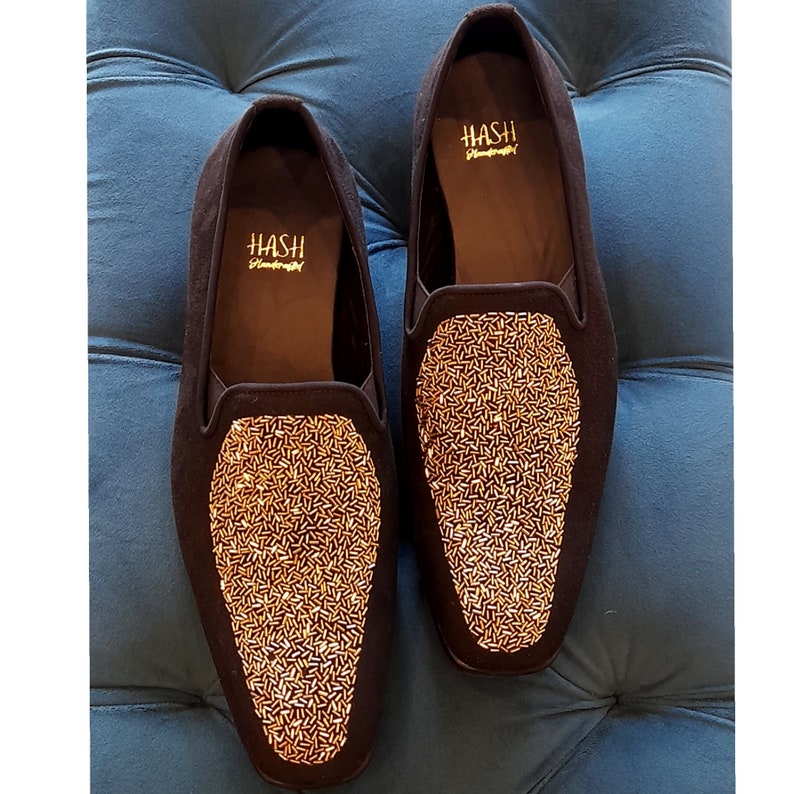 May include: A pair of black velvet loafers with gold beaded embellishments. The loafers have a pointed toe and a slip-on design. The brand name "HASH" is printed on the inside of each shoe.