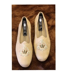 May include: A pair of beige suede slippers with gold and red embroidery. The embroidery features a floral design with a central motif of two stylized tulips.