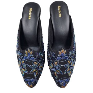 May include: A pair of black embroidered slippers with blue and gold floral designs. The slippers have a pointed toe and a low heel. The brand name "Shrivas" is printed on the insole.