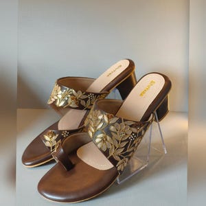 May include: A pair of brown block-heeled sandals featuring a toe-post design and a wide strap across the top. The straps are decorated with intricate gold and cream floral embroidery. The brand name Shrivas is printed on the inner sole.