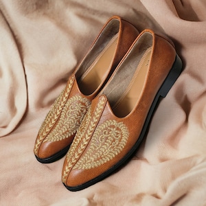 May include: A pair of brown leather loafers with gold embroidery. The shoes have a classic design with a rounded toe and a low heel. The loafers are displayed on a soft, neutral-toned fabric. The shoes are made of leather.