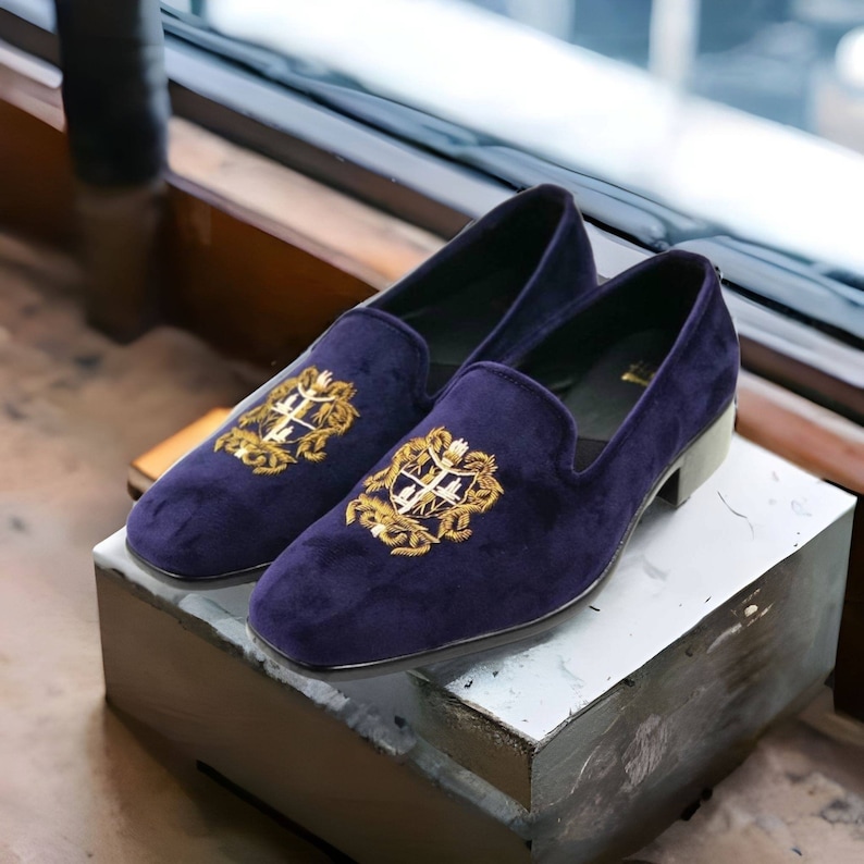 Puede incluir: Un par de zapatillas de terciopelo morado oscuro con crestas bordadas en oro. Las zapatillas tienen una punta puntiaguda y un tac&oacute;n bajo.