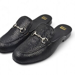 Puede incluir: Un par de mocasines de cuero negro con estampado de piel de serpiente y una hebilla de metal plateado. Los mocasines son de estilo deslizante y tienen una punta redonda.