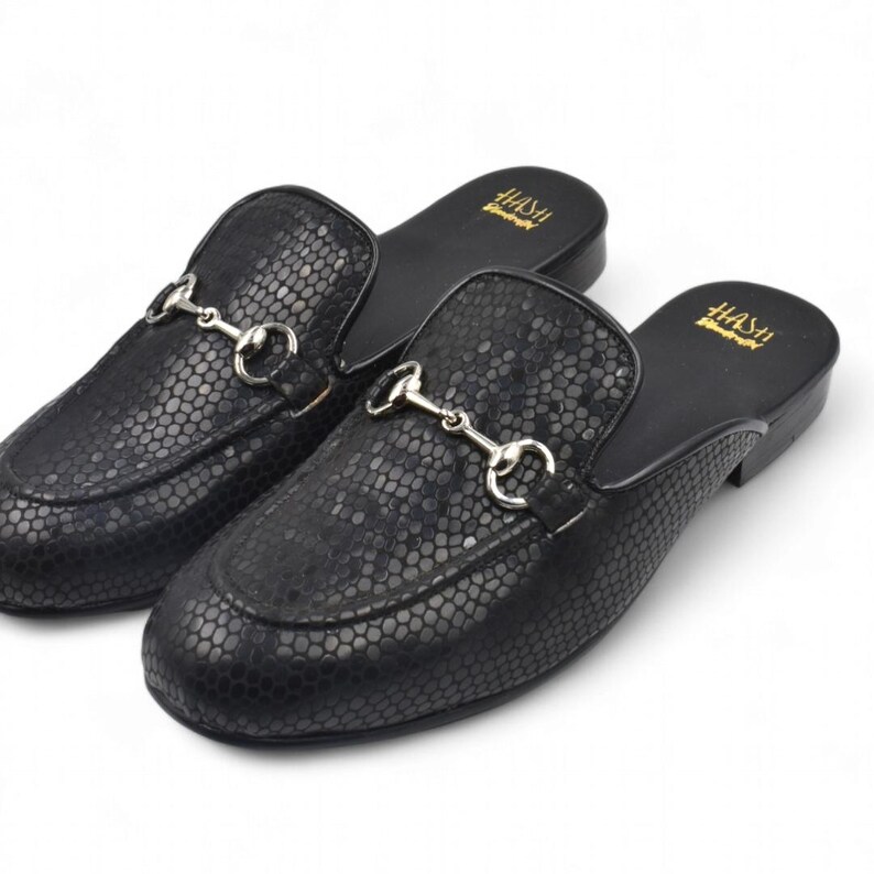 Puede incluir: Un par de mocasines de cuero negro con estampado de piel de serpiente y una hebilla de metal plateado. Los mocasines son de estilo deslizante y tienen una punta redonda.