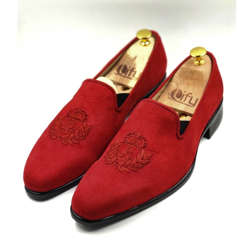 May include: A pair of red velvet loafers with a black sole and a gold embroidered crest on the toe.