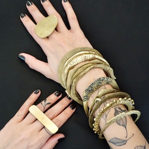 Op de afbeelding: Twee handen dragen meerdere gouden armbanden en ringen. De armbanden zijn gestapeld en hebben een gehamerde textuur. De ringen zijn eenvoudig en hebben een gladde afwerking.