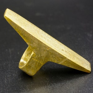 May include: A gold-colored metal ring with a unique, angular design. The ring has a flat, wide band that extends out to form a sharp point.