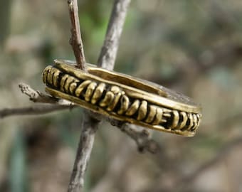 May include: A gold-colored ring with a decorative, textured band. The band is adorned with a repeating pattern of raised, geometric shapes.