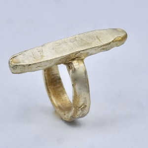 May include: A gold-colored ring with a rectangular top. The ring has a textured, hammered appearance, giving it a rustic, handcrafted look. The band is a simple, rounded design. The ring is likely made of metal.