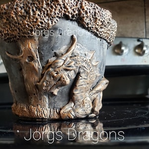 May include: A gray concrete planter with a dragon and tree design. The dragon is gold and the tree is gold with a rough texture. The planter has the words "Jorgs Dragons" written on it.