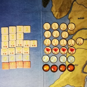 Puede incluir: Un tablero de juego con una variedad de fichas y dados de colores. Las fichas est&aacute;n numeradas del 0 al 3 y tienen un ojo rojo en el centro. Los dados est&aacute;n numerados del 1 al 3 y son amarillos con n&uacute;meros negros. El tablero est&aacute; dividido en secciones con diferentes nombres, incluyendo "Enedwaith" y "Andrast".