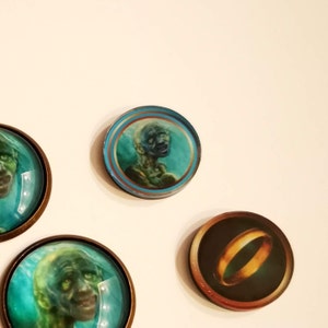 Puede incluir: Cuatro cabujones redondos de vidrio con marcos de metal. Los cabujones presentan im&aacute;genes de un anillo y tres caras diferentes. Las caras son verdes y azules, y el anillo es dorado.