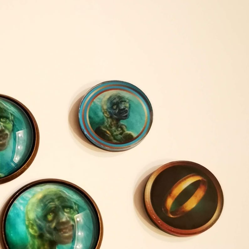 Puede incluir: Cuatro cabujones redondos de vidrio con marcos de metal. Los cabujones presentan im&aacute;genes de un anillo y tres caras diferentes. Las caras son verdes y azules, y el anillo es dorado.