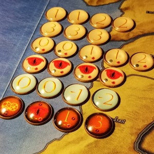 May include: A collection of 20 round game tokens with various numbers and symbols. The tokens are gold, white, and red with black outlines. The numbers include 0, 1, 2, 3, and -1. The symbols include an eye with a red pupil.