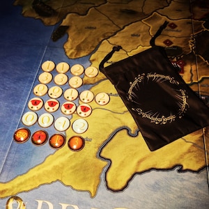 May include: A brown cloth bag with the inscription "One Ring to rule them all, One Ring to find them, One Ring to bring them all and in the darkness bind them" and a set of 20 round tokens with numbers and red and yellow symbols.