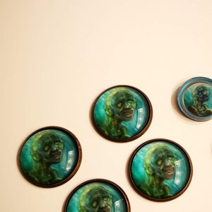 May include: Five round, glass cabochons with bronze metal settings. Each cabochon features a green and blue image of a creature with a human-like face.
