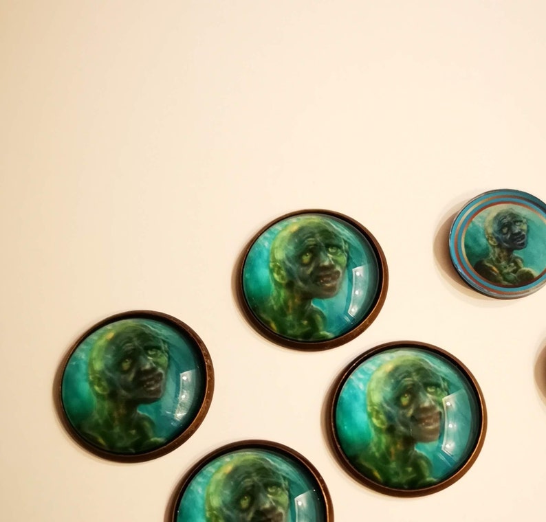 May include: Five round, glass cabochons with bronze metal settings. Each cabochon features a green and blue image of a creature with a human-like face.