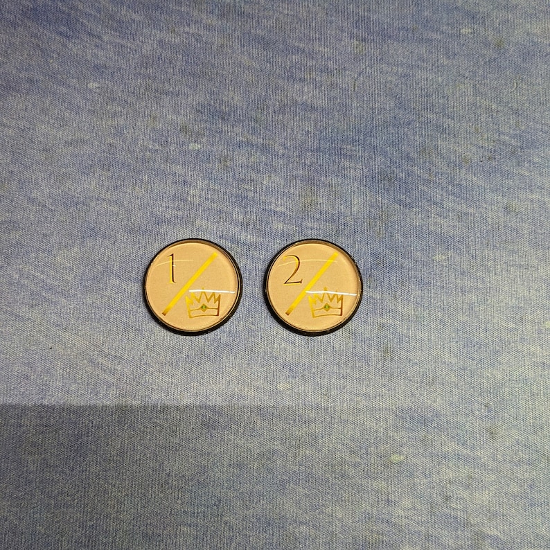 May include: Two round, beige-colored tokens with black borders. The tokens have a gold crown and a diagonal line with the numbers "1" and "2" on each.