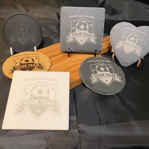 May include: A selection of coasters in different shapes, including round, square, and heart-shaped. Each coaster displays a football design with the text "YOUR TEAM" and "YOUR CLUB LOGO HERE". The coasters are made from materials such as slate and cork.
