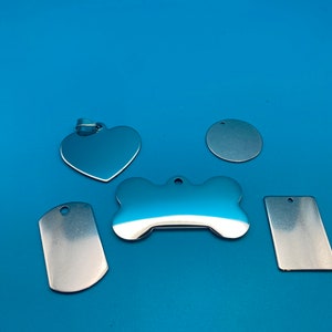 May include: A selection of polished, silver pet ID tags in different forms: a heart, a circle, a bone, a military-style tag, and a rectangular tag. Each tag has a small hole for attaching to a collar. The tags are set against a vibrant blue backdrop.