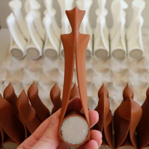 Op de afbeelding: Een terracotta-kleurige flesstopper met een uniek ontwerp, vastgehouden in een hand. De stopper heeft een cirkelvormige basis en een decoratieve bovenkant. Andere stoppers zijn zichtbaar op de achtergrond, sommige in het wit.