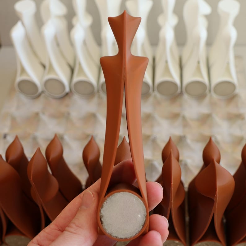 Op de afbeelding: Een terracotta-kleurige flesstopper met een uniek ontwerp, vastgehouden in een hand. De stopper heeft een cirkelvormige basis en een decoratieve bovenkant. Andere stoppers zijn zichtbaar op de achtergrond, sommige in het wit.