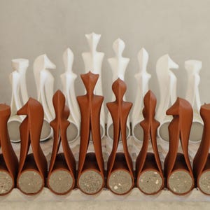 Op de afbeelding: Een schaakspel met terracotta en witte stukken. De terracotta stukken hebben een uniek, gestileerd ontwerp met een cilindrische basis. De witte stukken hebben een abstracte, langwerpige vorm. De set wordt gepresenteerd op een lichtgekleurd oppervlak.