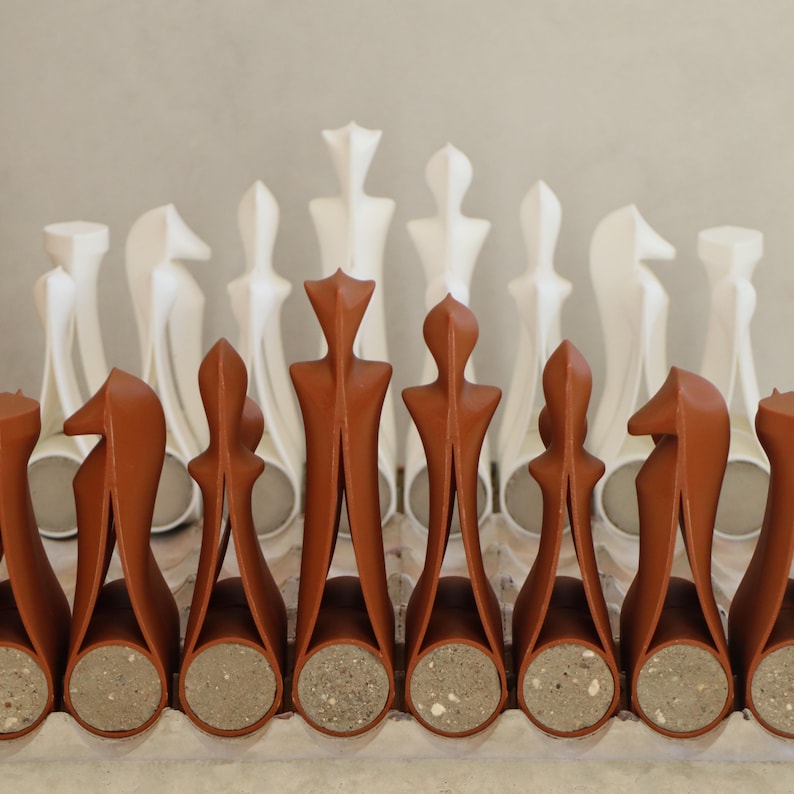 Op de afbeelding: Een schaakspel met terracotta en witte stukken. De terracotta stukken hebben een uniek, gestileerd ontwerp met een cilindrische basis. De witte stukken hebben een abstracte, langwerpige vorm. De set wordt gepresenteerd op een lichtgekleurd oppervlak.