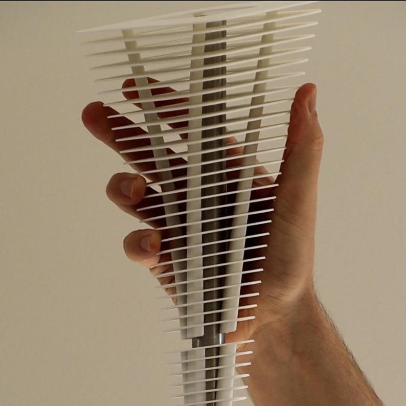 May include: A white, abstract sculpture composed of numerous horizontal, thin, rectangular pieces. The sculpture is held by a hand, showcasing its layered design. The artwork appears to be made of plastic or a similar material, with a minimalist aesthetic.
