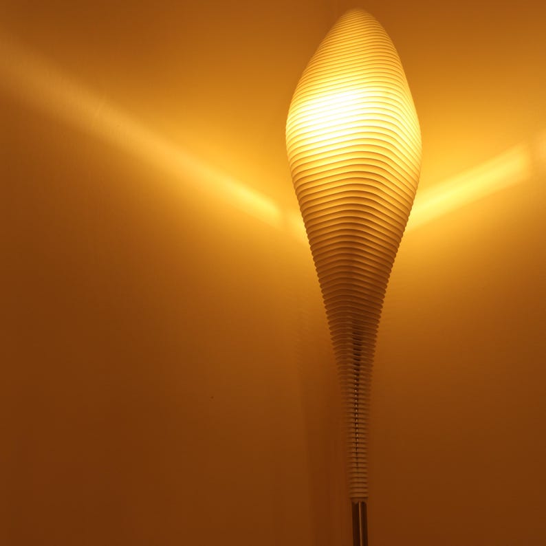May include: A tall, modern floor lamp with a unique, layered design. The lamp has a long, slender shape with a series of horizontal, curved elements that create a textured surface. The lamp emits a warm, inviting glow, perfect for ambient lighting.