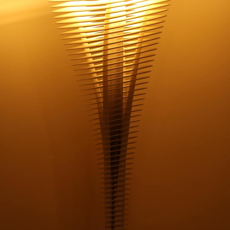 May include: A modern, abstract lamp with a layered design. The lamp features numerous horizontal wooden slats that create a conical shape. The warm light emanating from within casts a soft glow against the amber background.