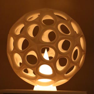 May include: A spherical, cream-coloured lamp with a geometric design. The lamp has numerous oval-shaped cutouts, allowing light to shine through. It sits on a white base, and the interior light source is visible. The overall design is modern and decorative.