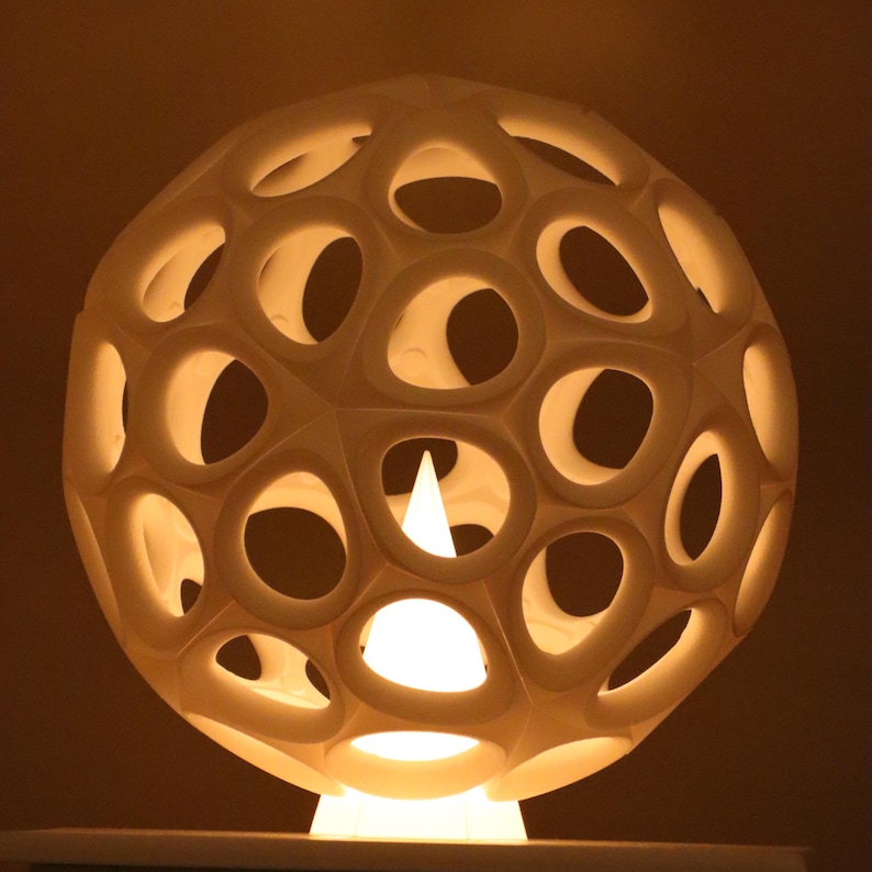 May include: A spherical, cream-coloured lamp with a geometric design. The lamp has numerous oval-shaped cutouts, allowing light to shine through. It sits on a white base, and the interior light source is visible. The overall design is modern and decorative.
