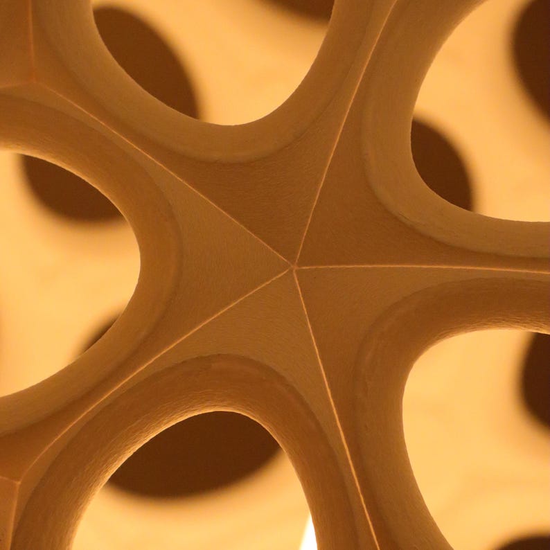 May include: Close-up of a light brown, geometric architectural detail. The design features a central star shape with curved, open sections radiating outwards. The structure has a smooth, matte finish and is illuminated with warm, ambient lighting.