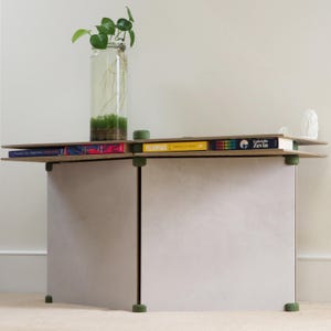 May include: A modern, two-tiered side table with a light gray base and a wooden top. The top shelf holds books and a small green object. A glass vase with a plant sits on top. The table is supported by green cylindrical feet.