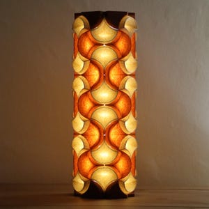 May include: A tall, rectangular lamp with a repeating pattern of orange and cream-colored curved shapes. The lamp has a dark brown wooden frame and emits a warm, inviting glow. The design is reminiscent of mid-century modern decor.