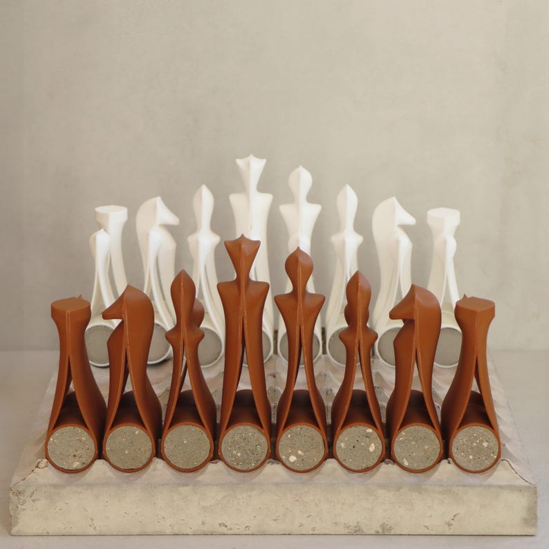Op de afbeelding: Een modern schaakspel met bruine en witte stukken. De bruine stukken hebben een uniek, abstract ontwerp, terwijl de witte stukken traditioneler zijn. De set wordt gepresenteerd op een betonnen bord, wat een minimalistische uitstraling cre&euml;ert.