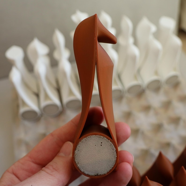 Op de afbeelding: Een bruine flesstopper met een uniek, gebogen ontwerp. De stopper heeft een cilindrische basis met een grijs, poreus oppervlak. Het ontwerp doet denken aan een muzieknoot. Andere witte decoratieve objecten zijn op de achtergrond.