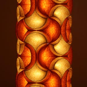 May include: A close-up of a decorative lamp with a repeating pattern of orange and gold shapes. The lamp has a warm, inviting glow, with the light filtering through the patterned design. The overall effect is one of texture and depth.