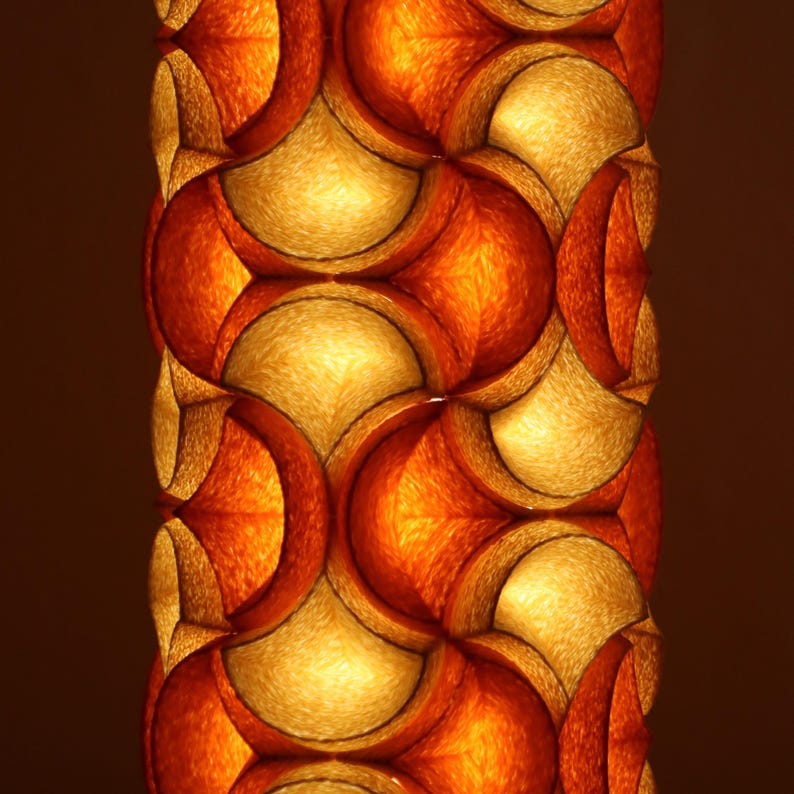 May include: A close-up of a decorative lamp with a repeating pattern of orange and gold shapes. The lamp has a warm, inviting glow, with the light filtering through the patterned design. The overall effect is one of texture and depth.