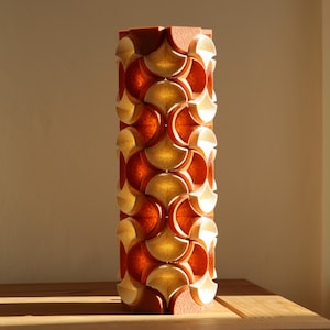 May include: A tall, decorative pillar with a repeating pattern of orange and gold fan-shaped cutouts. The pillar is made of a reddish-brown material and stands on a wooden surface. The design creates a play of light and shadow.
