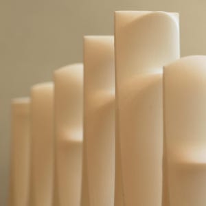 Op de afbeelding: Close-up van een rij cr&egrave;mekleurige, rechthoekige kaarsen. Elke kaars heeft een gebogen bovenkant en werpt zachte schaduwen. De achtergrond is een gedempte, neutrale kleur. De kaarsen zijn waarschijnlijk van was.