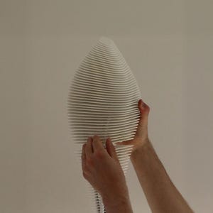 May include: A white, abstract sculpture with a layered, ribbed design. The sculpture has a teardrop shape and is held by two hands. The background is a plain, off-white wall. The sculpture's dimensions are not visible.