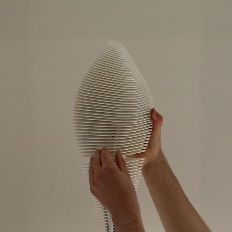 May include: A white, abstract sculpture with a layered, ribbed design. The sculpture has a teardrop shape and is held by two hands. The background is a plain, off-white wall. The sculpture's dimensions are not visible.