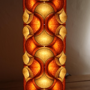 May include: A tall, cylindrical lamp with a repeating pattern of orange and gold geometric shapes. The lamp is illuminated from within, creating a warm, inviting glow. The design is reminiscent of mid-century modern decor.