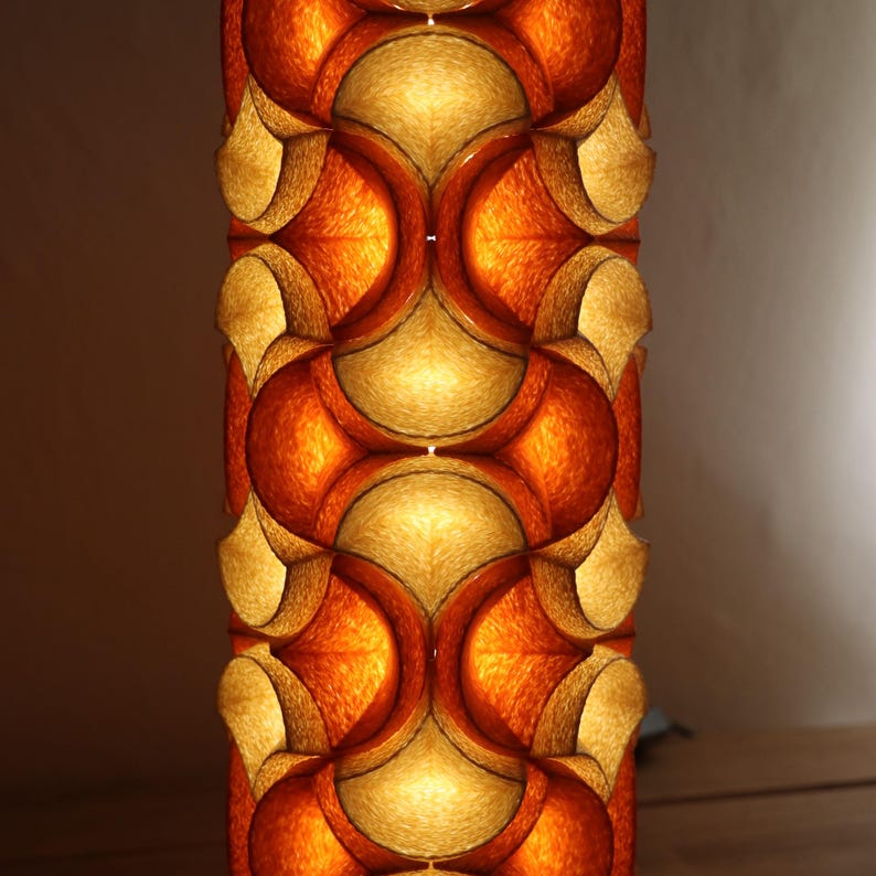 May include: A tall, cylindrical lamp with a repeating pattern of orange and gold geometric shapes. The lamp is illuminated from within, creating a warm, inviting glow. The design is reminiscent of mid-century modern decor.